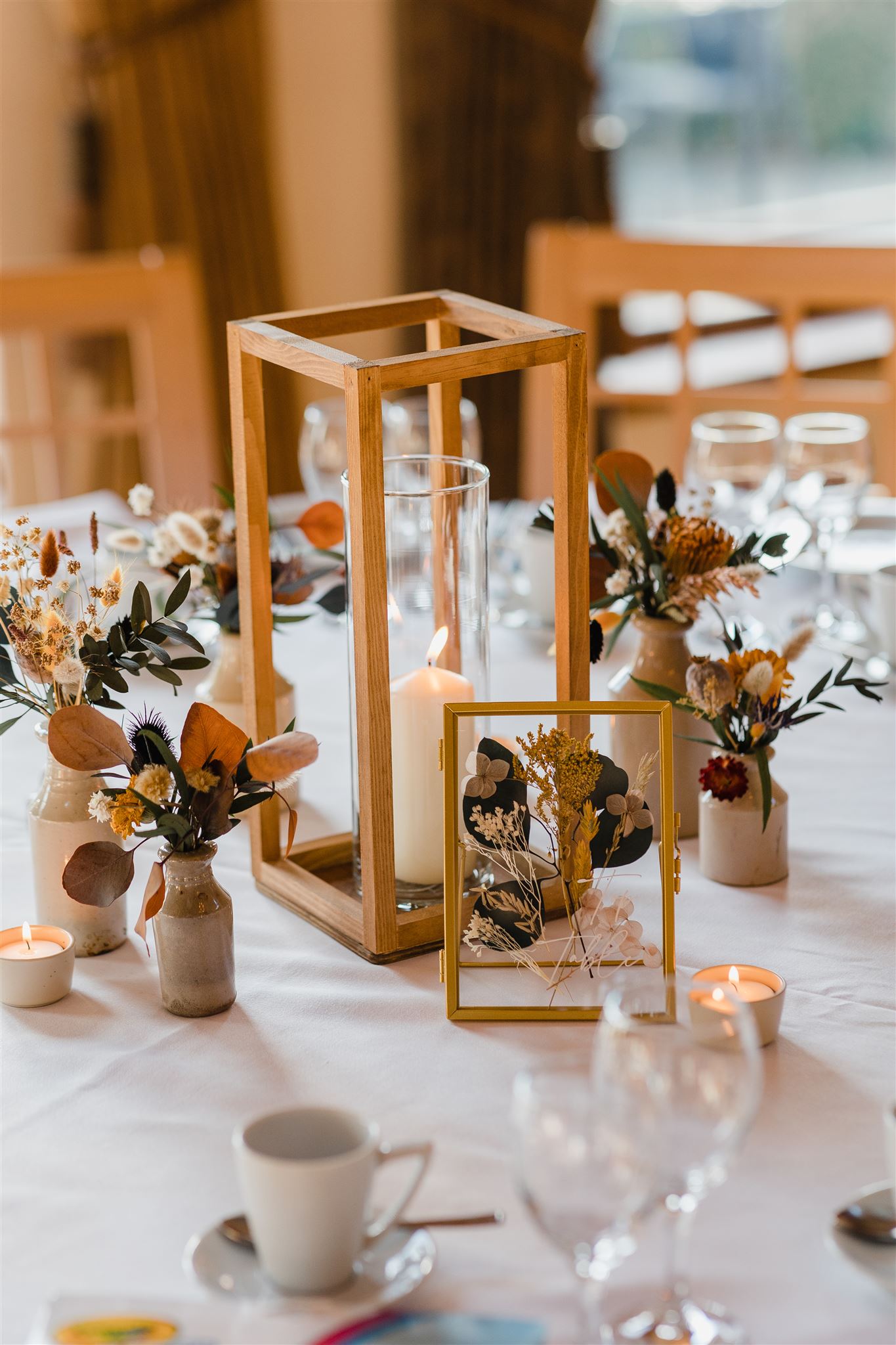 Dried flowers in small stoneware vases and a pillar candle in wooden box frame centre a wedding reception table.